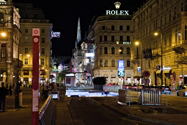 Eine nächtliche Stadtstraße mit fahrenden Autos, Fußgängern auf den Gehwegen, parkenden Fahrrädern, Straßenlaternen, hohen Gebäuden, Absperrungen, Pfosten und einem Rolex-Logo auf einem Gebäude im Hintergrund.