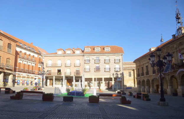 Ein Stadtplatz mit einem zentralen Brunnen, umgeben von Bänken, Topfpflanzen, Laternenmasten, Straßenlaternen und einem Uhrenturm im Hintergrund unter einem klaren blauen Himmel.
