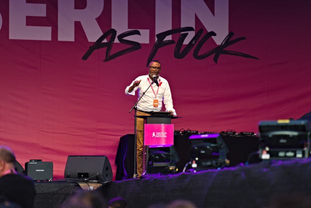 Ein Mann in einem weißen Hemd und braunen Hosen steht auf einer Bühne und hält ein Mikrofon, während ein pinker Banner mit der Aufschrift 'Berlin as Fuck' hinter ihm hängt.
