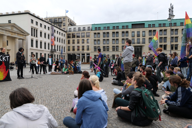Eine Gruppe von Menschen, die auf dem Boden sitzen, vor einer Menge mit Fahnen und Spruchbändern bei einer Anti-Schwulen-Demonstration in Berlin, mit einer Statue, Gebäuden und einem Mikrofonständer zu sehen.