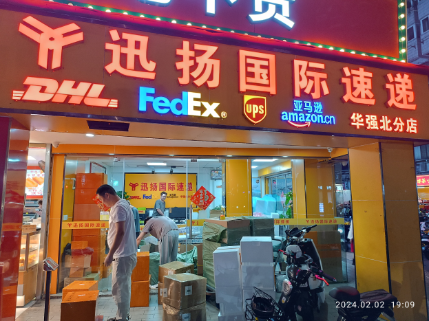 Ein DHL- und FedEx-Laden in Shanghai, China, mit Menschen, Kartons und Fahrzeugen davor und einem beleuchteten Gebäude im Hintergrund.