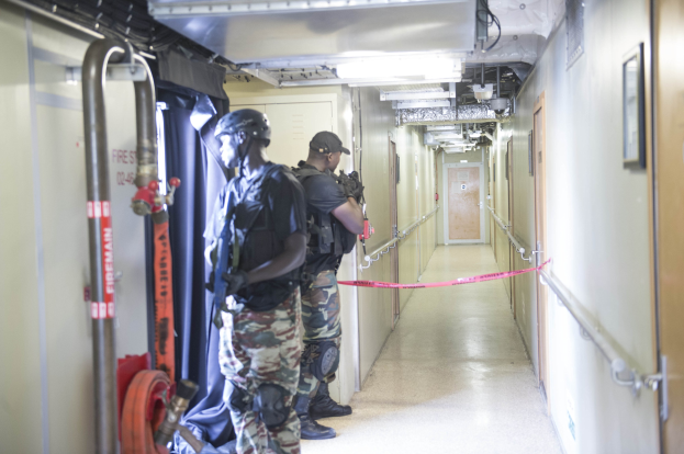 Zwei Polizisten in Uniform stehen nebeneinander in einem Flur, jeder trägt einen Helm und hat eine Waffe dabei, mit einer Tür und einem gerahmten Foto auf der rechten Wand und Deckenleuchten und Rohren.