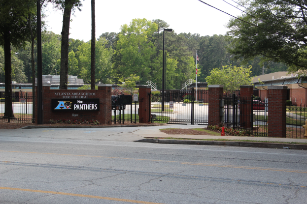 Der Eingang einer Schule mit einem Schild an der Seite, umgeben von Bäumen, Straßenlaternen, Gittern, Namensschildern, Kraftfahrzeugen auf der Straße, Verkehrskegeln, Pflanzen, Gras, Gebäuden und einem klaren blauen Himmel.