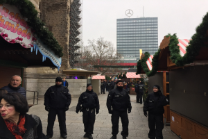 Polizeibeamte in schwarzen Uniformen stehen vor einem geschmützten Weihnachtsmarkt in Berlin mit festlichen Deko, Gebäuden, Bäumen und einem klaren blauen Himmel im Hintergrund.