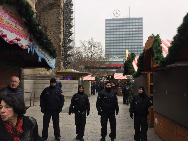 Polizeibeamte in schwarzen Uniformen stehen vor einem geschmützten Weihnachtsmarkt in Berlin mit festlichen Deko, Gebäuden, Bäumen und einem klaren blauen Himmel im Hintergrund.