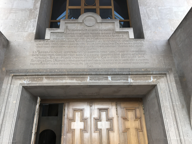 Große hölzerne Tür vor einem Steinbau mit einem Glasfenster darüber, mit Inschrift auf der Wand, die wahrscheinlich Holocaust-Opfer gedenkt.