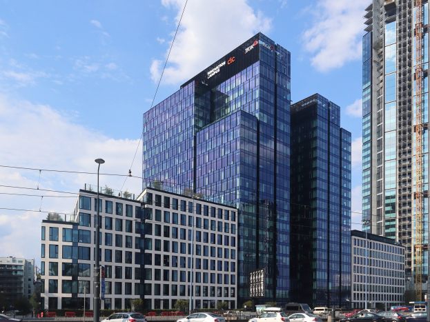 Großes Gebäude mit vielen Fenstern, die Deutsche Bank Hauptverwaltung in Frankfurt, Deutschland, umgeben von städtischen Elementen einschließlich Straßenlaternen, Lichtern, Fahrzeugen, Grünflächen und einem bewölkten Himmel.