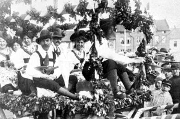 Schwarzes Bild von Menschen in traditioneller bayrischer Tracht auf einem geschmückten Paradewagen, einige spielen Musikinstrumente, mit Gebäuden und einem klaren Himmel im Hintergrund.