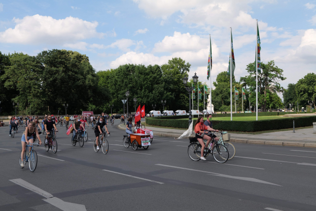 Eine Gruppe von Menschen, die auf Fahrrädern eine von Bäumen gesäumte Straße mit Laternenmasten und Flaggenmasten entlangfahren, während der Pariser Fahrradtour 2016, mit bewölktem Himmel und ein paar Fahrzeugen im Hintergrund.