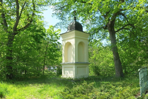 Kleines weißes Holocaust-Denkmal auf einem grasbewachsenen Friedhof umgeben von einem Zaun und Bäumen unter einem klaren blauen Himmel in Vilnius, Litauen.