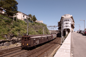 Güterzug fährt auf Schienen neben einem Gebäude mit parkenden Autos auf einem Bahnsteig zu seiner Rechten, vor einem Hintergrund aus städtischen Strukturen, Bäumen, Strommasten, Drähten und einem klaren blauen Himmel.