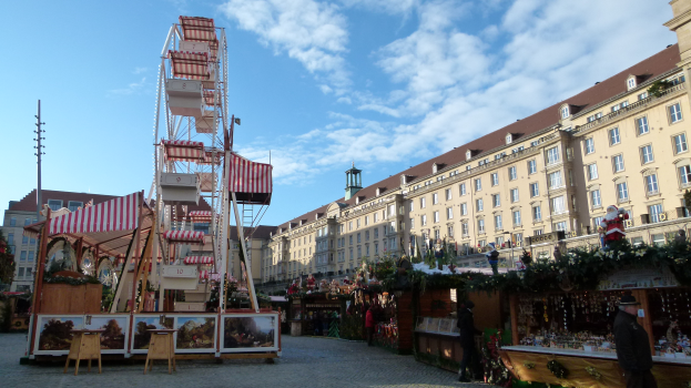 Ein lebhafter Weihnachtsmarkt auf einem Stadtplatz, mit einem zentralen Riesenrad, umgeben von Gebäuden, Bäumen und einem bewölkten Himmel, mit zahlreichen Menschen und verschiedenen Gegenständen, die über das Gebiet verstreut sind.