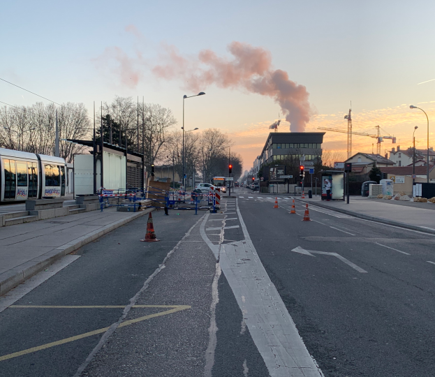 Eine Stadtstraße mit Verkehrskegeln, einem Bus, Laternen, Bäumen, Gebäuden, Kränen und Rauch, der aus einem entfernten Gebäude aufsteigt.