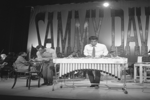 Eine Gruppe von Männern, die auf einer Bühne Marimbas spielen, beleuchtet von Scheinwerfern und umgeben von einem Banner mit Text, in Schwarz-Weiß.