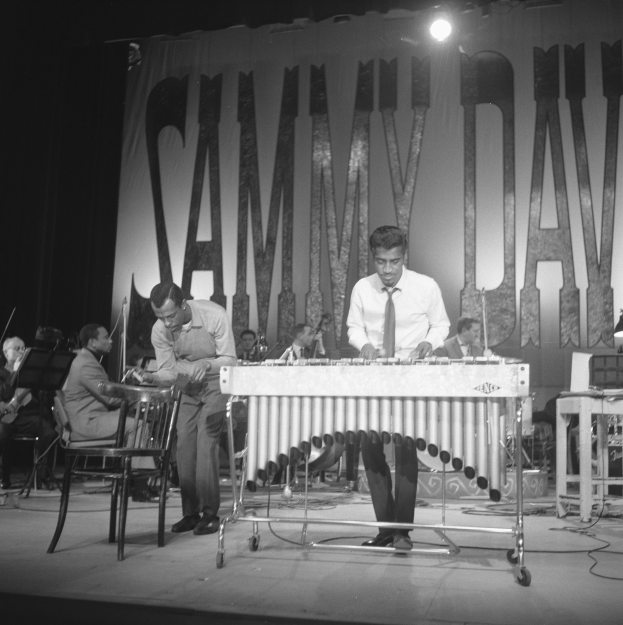 Eine Gruppe von Männern, die auf einer Bühne Marimbas spielen, beleuchtet von Scheinwerfern und umgeben von einem Banner mit Text, in Schwarz-Weiß.