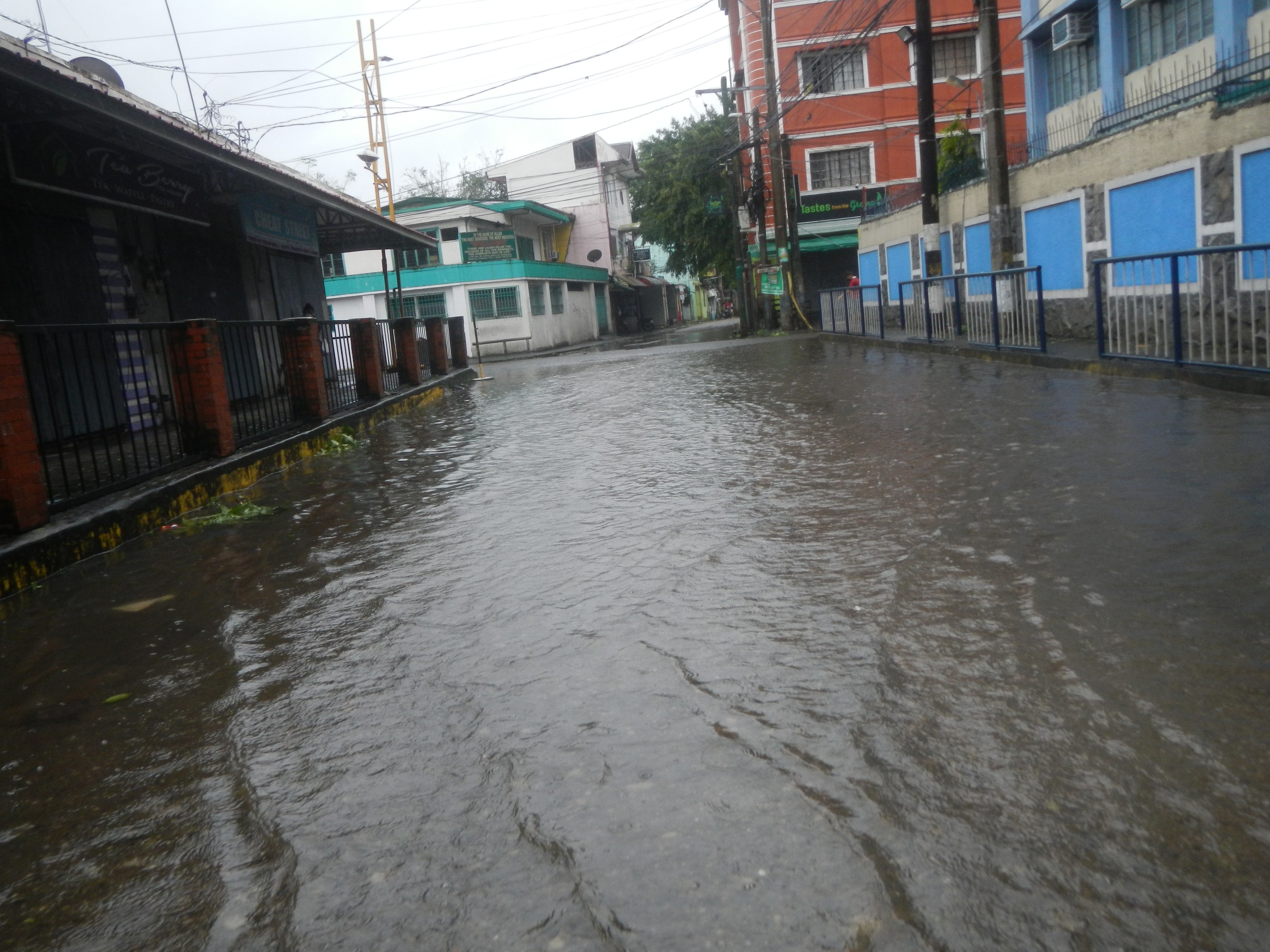 Flutstraßen in der Stadt mit Wasser bedeckt, das die Straße, Geländer, Gebäude, Strommasten, Bäume und den Himmel sichtbar macht.
