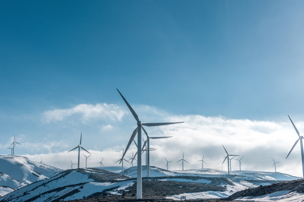 Eine Gruppe von Windkraftanlagen in einem verschneiten Feld mit verschneiten Hügeln und Wolken im Hintergrund, wahrscheinlich Teil eines Erneuerbare-Energien-Projekts.