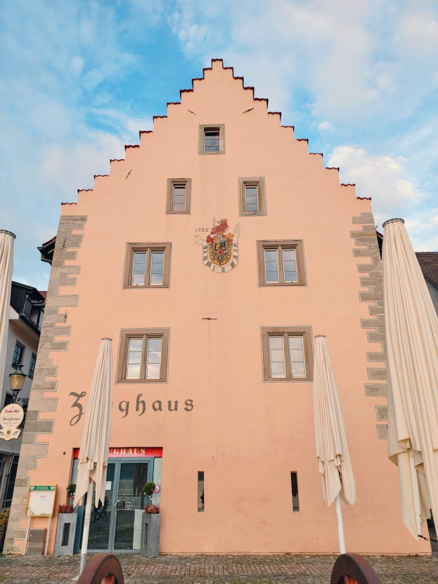 Pinkes Gebäude mit einem zentralen Uhrenturm, umgeben von Fenstern, Schirmen, einem Schild, einer Straßenlaterne, Topfpflanzen und einem bewölkten Himmel in München, Deutschland.