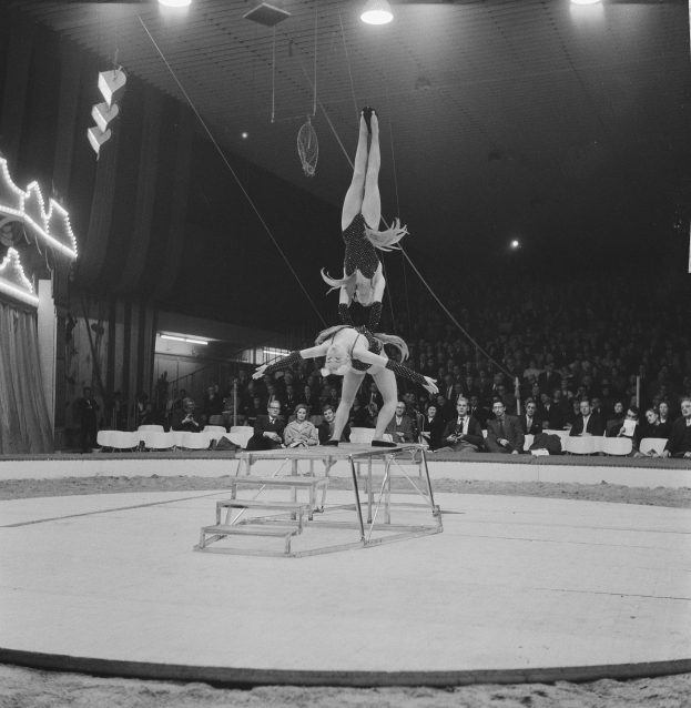 Zwei Artistinnen performen auf einer von Deckenleuchten beleuchteten Bühne, mit einem sitzenden Publikum im Hintergrund.