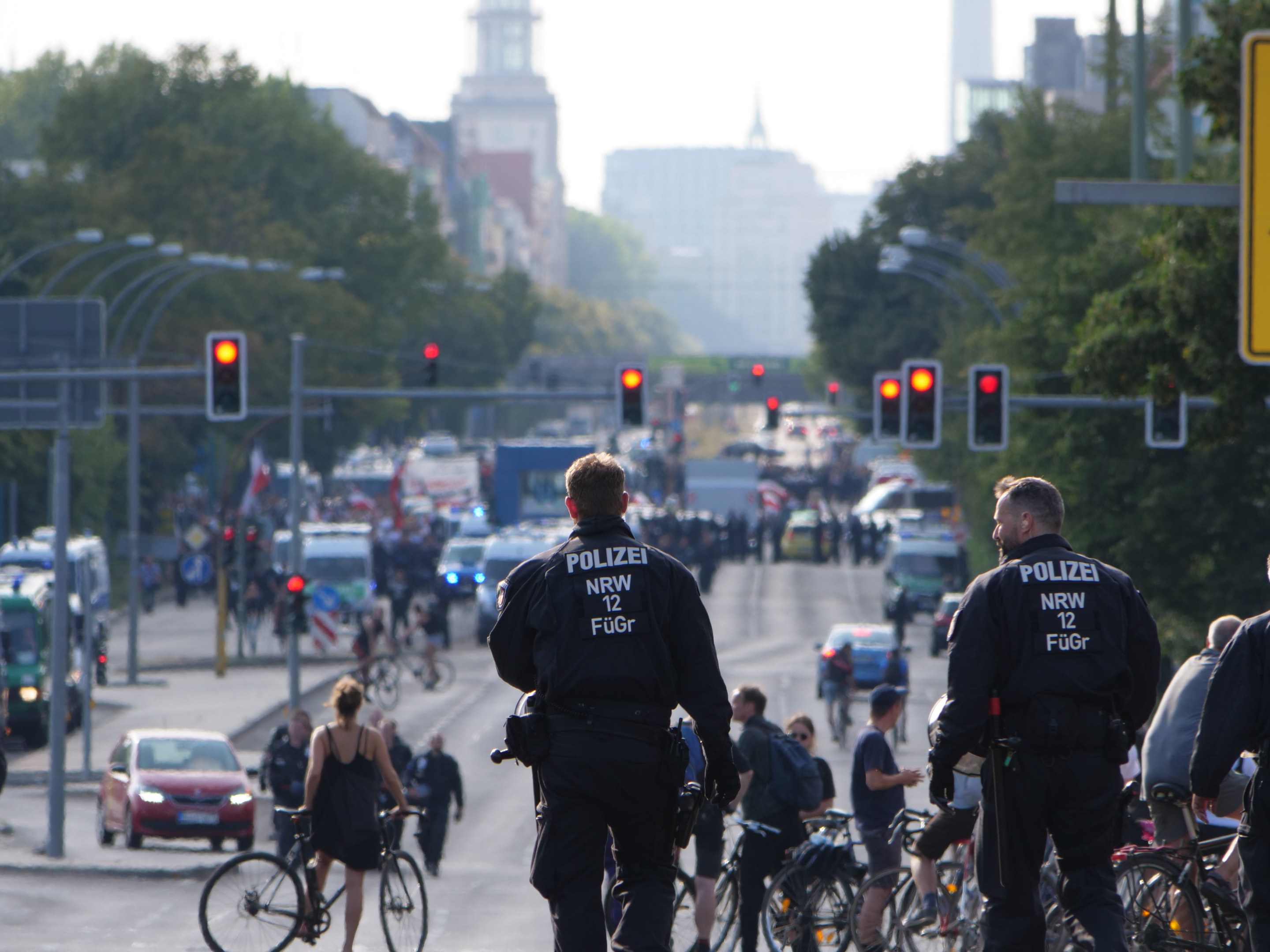 Polizeibeamte fahren mit Fahrrädern eine von Bäumen gesäumte Straße entlang mit Gebäuden und einem klaren blauen Himmel im Hintergrund.
