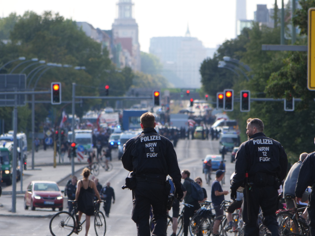 Polizeibeamte fahren mit Fahrrädern eine von Bäumen gesäumte Straße entlang mit Gebäuden und einem klaren blauen Himmel im Hintergrund.