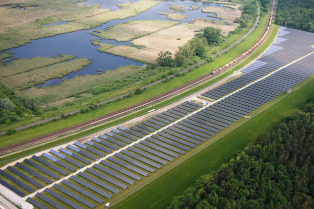 Luftaufnahme eines Solarparks mit Panelen in einem Feld, umgeben von Bäumen, Gras, Pflanzen und Wasser, mit einem Zug auf einer näheren Eisenbahnschiene.