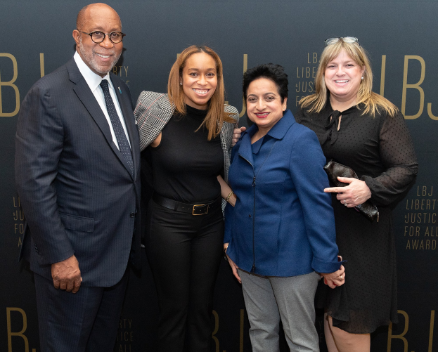 Eine Gruppe von vier Menschen auf einem roten Teppich, die lächeln und für ein Foto posieren, mit einem 'Liberty Justice for All Awards'-Schild im Hintergrund.
