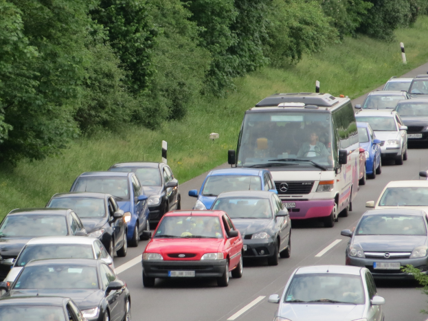 Ein Stau auf einer Autobahn mit vielen Autos und einem Lieferwagen, Menschen in den Fahrzeugen, mit Bäumen und Gras im Hintergrund.