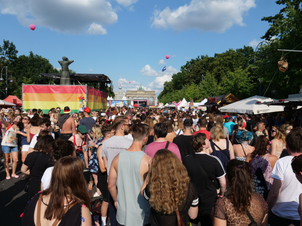 Eine große Menschenmenge, die eine Straße mit Zelten, Bäumen, Pfählen, Lichtern und einer Statue entlanggeht, mit Gebäuden und einem Himmel voller Wolken und Ballons im Hintergrund während des Christopher Street Day in Berlin.