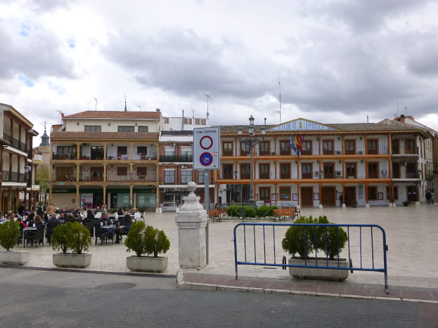 Ein belebter Stadtplatz mit Menschen, die sitzen und stehen, Topfpflanzen, Metallabsperrungen, Straßenlaternen mit Flaggen, ein Schild, Gebäude mit Fenstern und einem bewölkten Himmel.