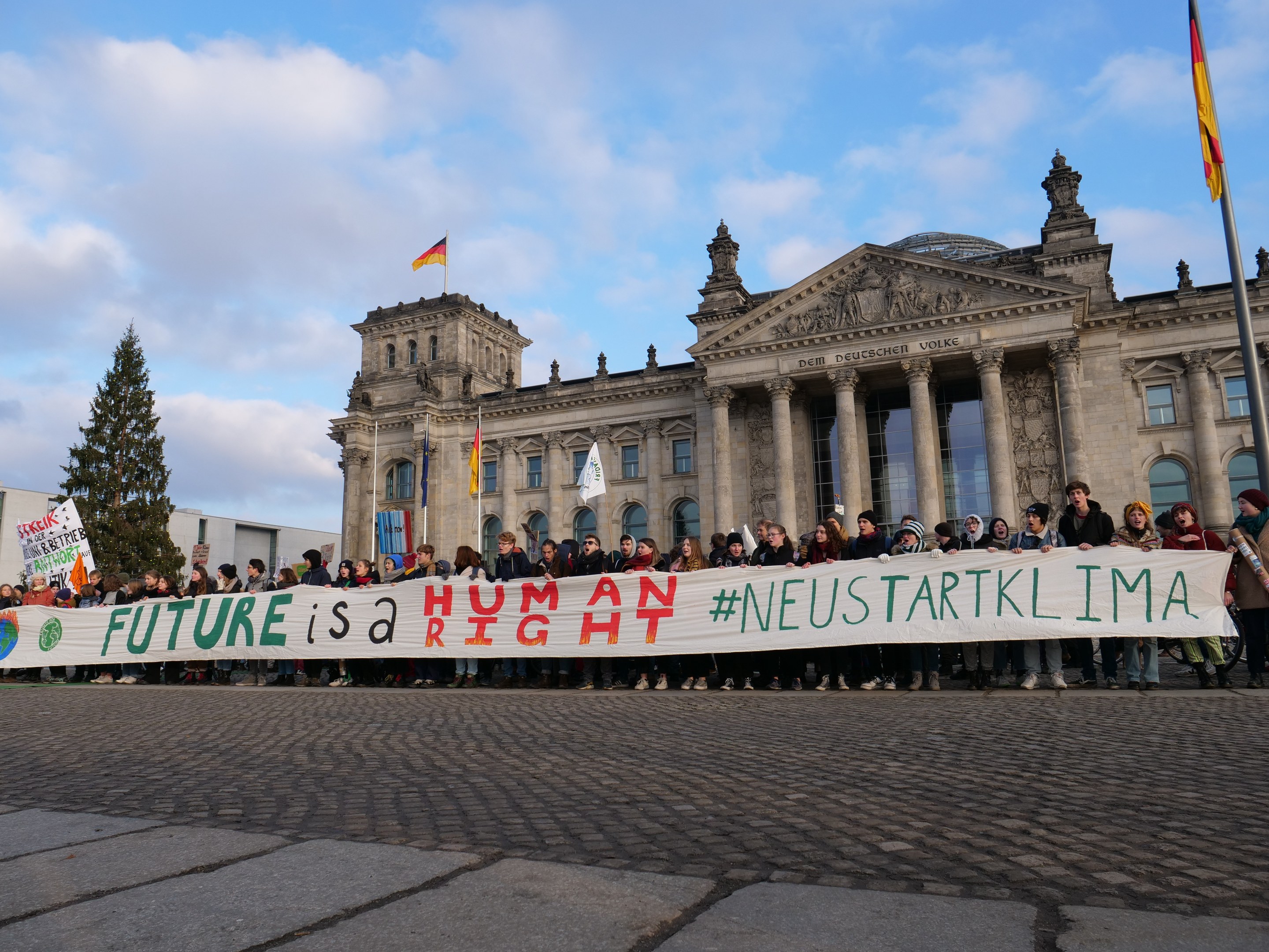 Gruppe von Menschen vor dem Reichstaggebäude in Berlin mit einer Fahne, auf der 'Zukunft ist ein Menschenrecht' steht