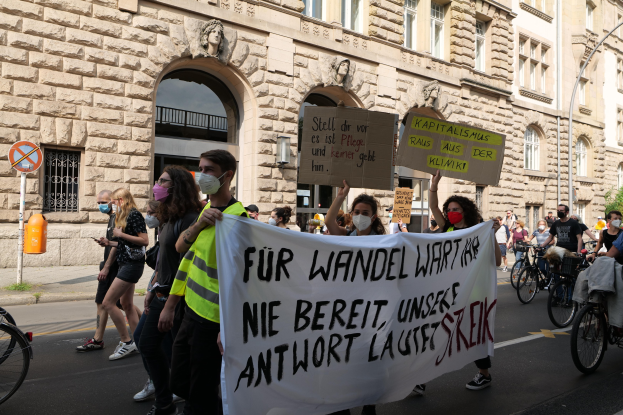 Eine Gruppe von Menschen marschiert durch eine Straße, einige halten Protestschilder und andere fahren Fahrräder, mit einem Gebäude im Hintergrund, das Fenster, Bögen, Säulen und Skulpturen aufweist.
