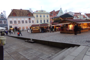 Ein lebendiger Weihnachtsmarkt in Tallinn, Estland, mit Menschen um geschmückte Stände, festliche Lichter und Gebäude mit Fenstern im Hintergrund, unter einem bewölkten Himmel, mit Stufen, die zum Markt führen, und Töpfen mit Pflanzen, die herumstehen.
