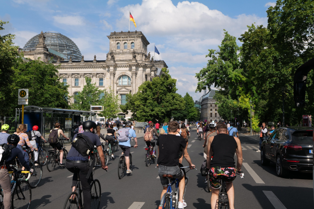 Eine Gruppe von Menschen, die auf Fahrrädern eine von Bäumen gesäumte Straße in Berlin entlangfahren, mit Gebäuden und einer Bushaltestelle im Hintergrund, unter einem bewölkten Himmel und einer Flagge auf einem der Gebäude.