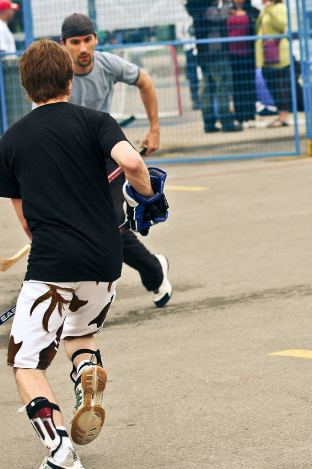 Zwei Personen, die Hockey auf einer Straße mit einem Zaun dahinter und einer Menge von Menschen im Hintergrund spielen.
