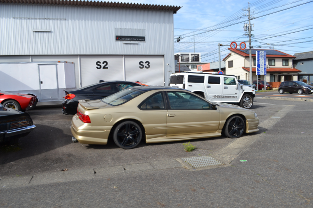Ein goldener Wagen auf einer Straße vor einem Gebäude geparkt, mit anderen Fahrzeugen und städtischen Elementen wie Polen, Drähten und Brettern im Hintergrund unter einem klaren blauen Himmel.