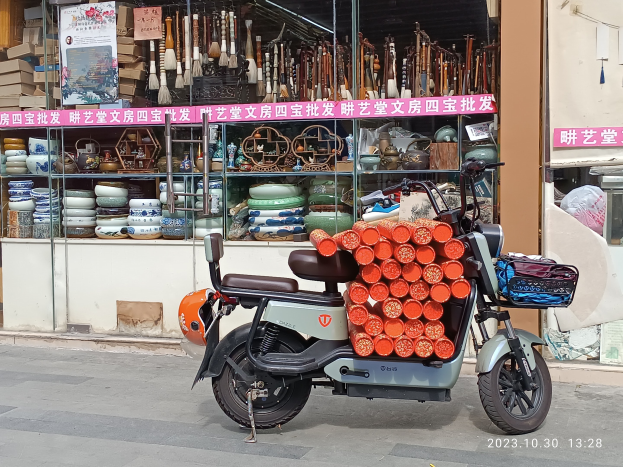 Ein Motorroller mit einem Korb ist auf der Straße vor einem Laden mit einer Glaswand geparkt, auf der Schalen, Teller und andere Gegenstände ausgestellt sind, mit einer sichtbaren Texttafel im Inneren.