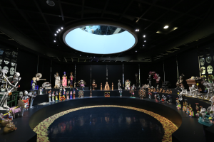 Ein großer Raum voller verschiedener Spielzeuge, darunter Skelette, die auf dem Boden angeordnet sind, mit Schädeln an den Wänden und Lichtern an der Decke, die eine festliche Atmosphäre für den Tag der Toten schaffen.