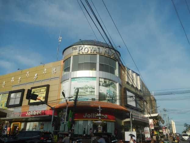 Eine belebte Straße mit fahrenden Autos, Menschen auf dem Gehweg und dem Royal Mall Einkaufszentrum im Hintergrund, umgeben von Bäumen und Strommästen unter einem klaren Himmel.
