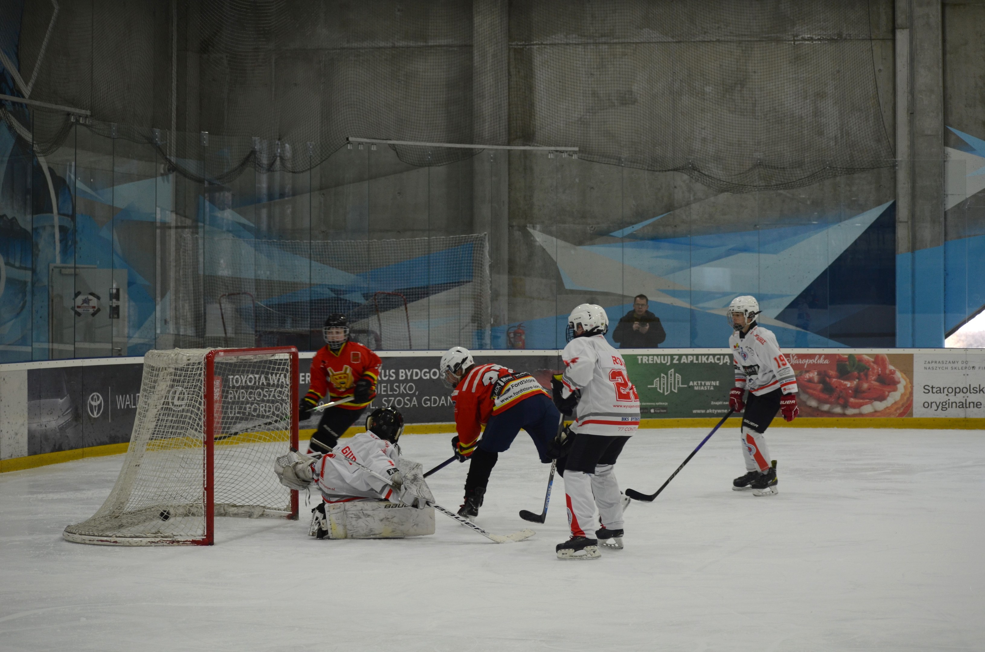 Eine Gruppe von Menschen, die auf einer Indoor-Eisbahn Eishockey spielen, tragen Helme und halten Hockeystöcke, mit einem Torpfosten links und Bannern im Hintergrund.