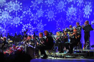 Eine Gruppe von Menschen, die auf einer Bühne Musikinstrumente spielen, mit Mikrofonen, Blumensträußen und einem mit Schneeflocken geschmückten Bildschirm im Hintergrund.