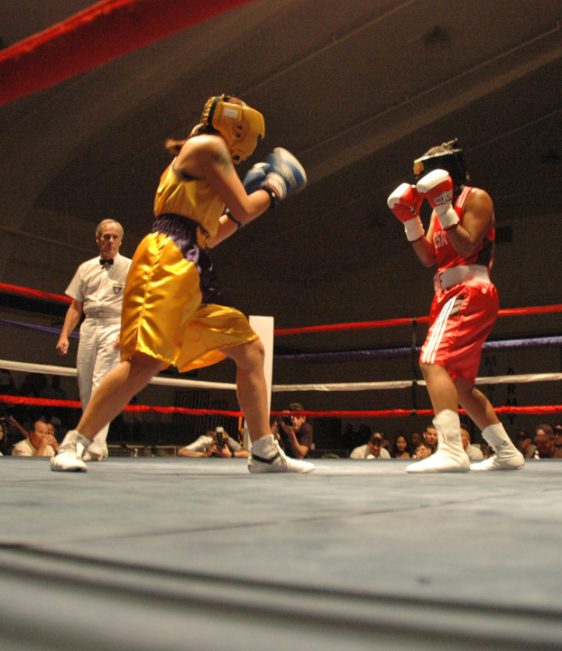 Zwei Boxer kämpfen im Ring, während ein Schiedsrichter beobachtet, umgeben von Fotografen und Zuschauern unter einer sichtbaren Decke.