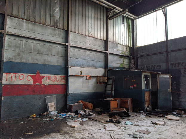 Verlassenes Gebäudeinneres mit Schutt, darunter Kartons, eine Leiter und verstreute Gegenstände, eine textbedeckte Wand links und Glasfenster rechts.