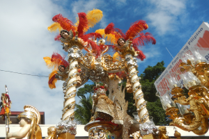 Ein mit goldenen und roten Gegenständen geschmückter Karnevalswagen, auf dem eine Gruppe von Menschen sitzt, mit einer Tafel auf der rechten Seite, vor einem Hintergrund aus Bäumen, einem Gebäude und einem bewölkten Himmel.