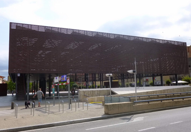 Großes modernes Gebäude mit zahlreichen Fenstern, das als neuer Eingang des Stadtzentrums dient, umgeben von Straßeninfrastruktur, Fahrzeugen, Fußgängern und Bäumen unter einem bewölkten Himmel.