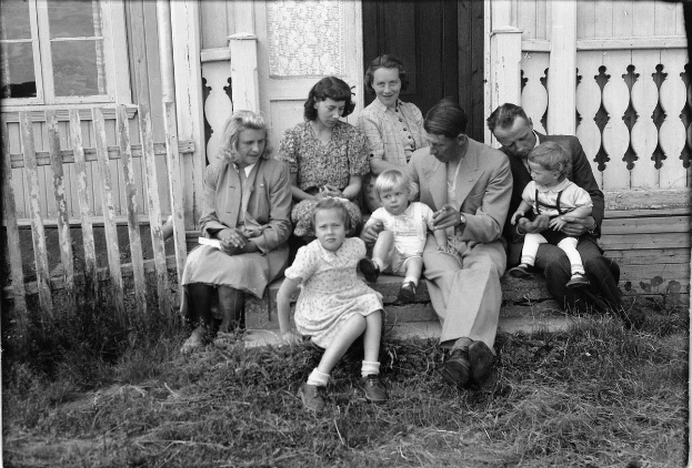 Ein Schwarz-Weiß-Foto einer Familie auf den Stufen eines Hauses, mit einigen sitzend und einigen stehend, einer hält ein Baby, mit einer Tür und einem Fenster im Hintergrund und Gras auf dem Boden.