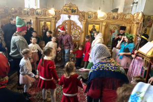 Eine Gruppe von Menschen steht vor einer Kirche, einige tragen Mützen, mit einem Buchstand rechts, Fotorahmen, Lichtern und einem Fenster im Hintergrund, während einer Weihnachtsfeier.