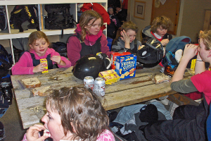 Eine Gruppe von Kindern sitzt um einen Esstisch und isst Essen, mit Taschen an einem Gestell darüber und Lebensmitteln auf dem Tisch.
