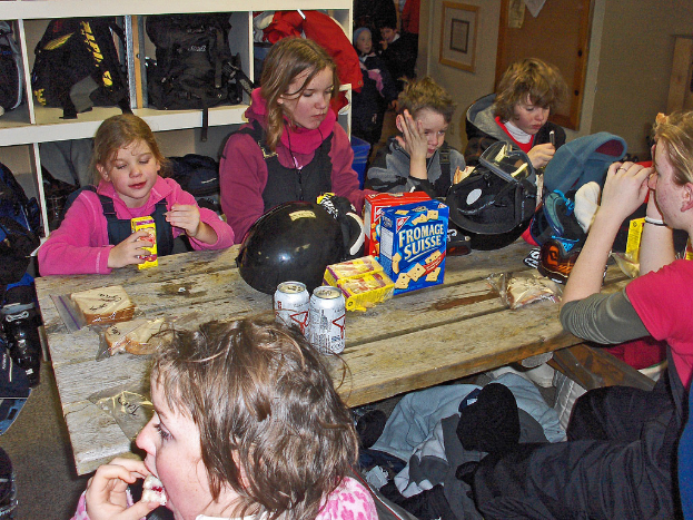 Eine Gruppe von Kindern sitzt um einen Esstisch und isst Essen, mit Taschen an einem Gestell darüber und Lebensmitteln auf dem Tisch.