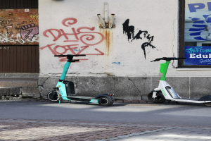 Zwei Elektro-Scooter nebeneinander auf einer Straße geparkt, mit einer Graffiti-bedeckten Wand und einem Plakat an einem Fenster im Hintergrund.
